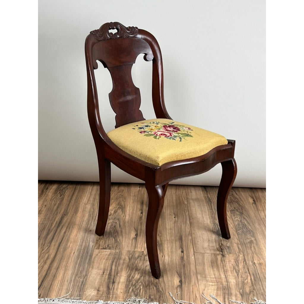 An Antique American Empire Flame Mahogany Saber Leg Side Chair (mid-19th century), featuring a carved backrest and floral-patterned yellow upholstery, is shown on a wood floor against a plain, light-colored wall.