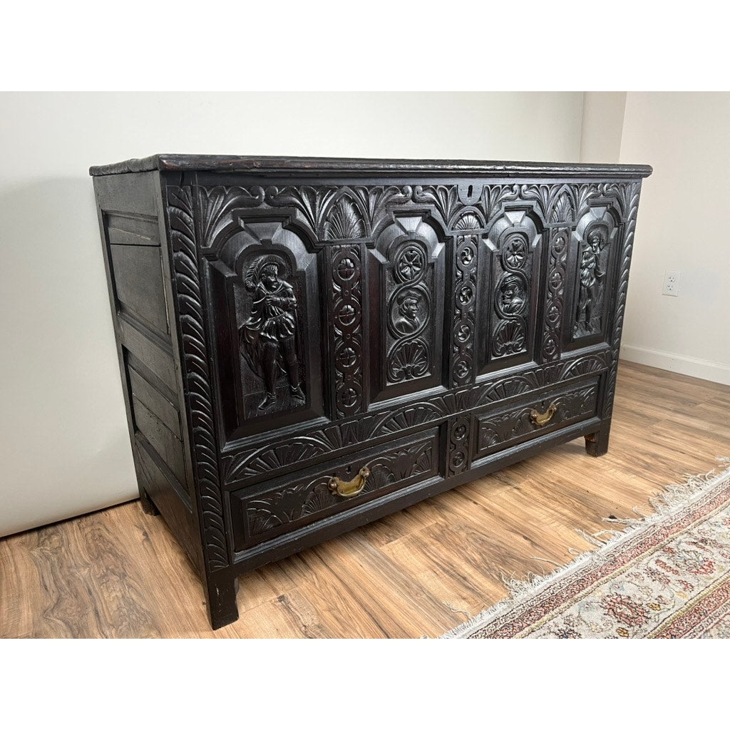 An Antique 18th Century Charles II Style Mule Chest, possibly Irish or Scottish, with ornate carvings and two bottom drawers, stands on a wood floor beside a patterned rug and a white wall. Brand unknown.