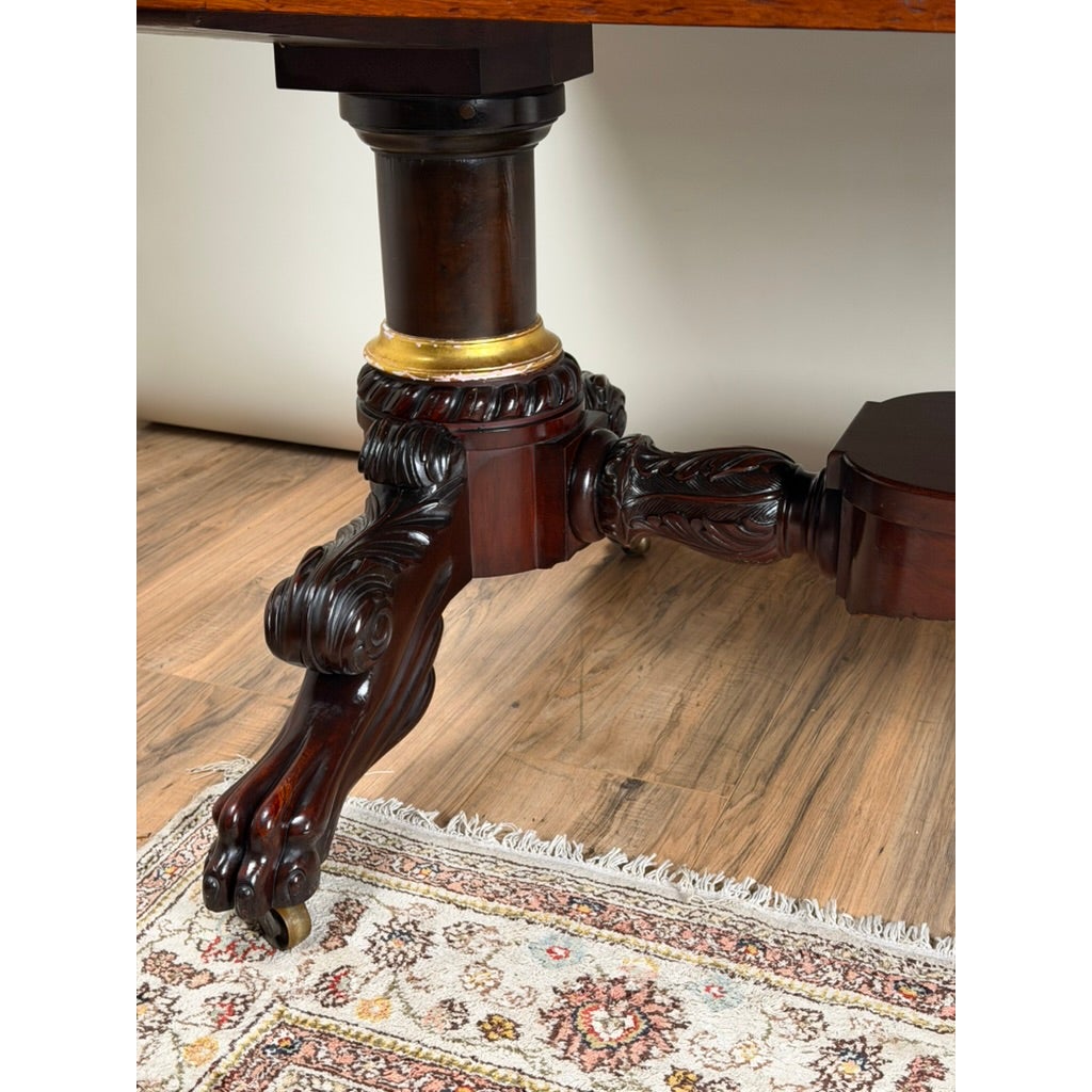 A mahogany leg on a New York Classical Revival Carved Mahogany Double-Pedestal Library Table, featuring ornate acanthus leaf carvings, a gilt-trimmed collar, and a claw-and-ball foot on a caster.