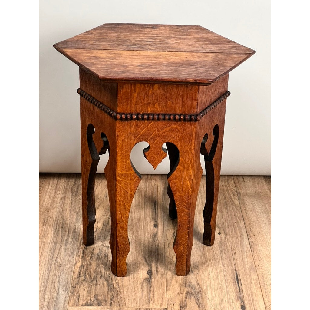 The Antique Arts & Crafts Small Oak Occasional Table c. 1910 features a hexagonal shape, carved legs, and decorative trim, shown here placed on a wooden floor against a plain light-colored wall.