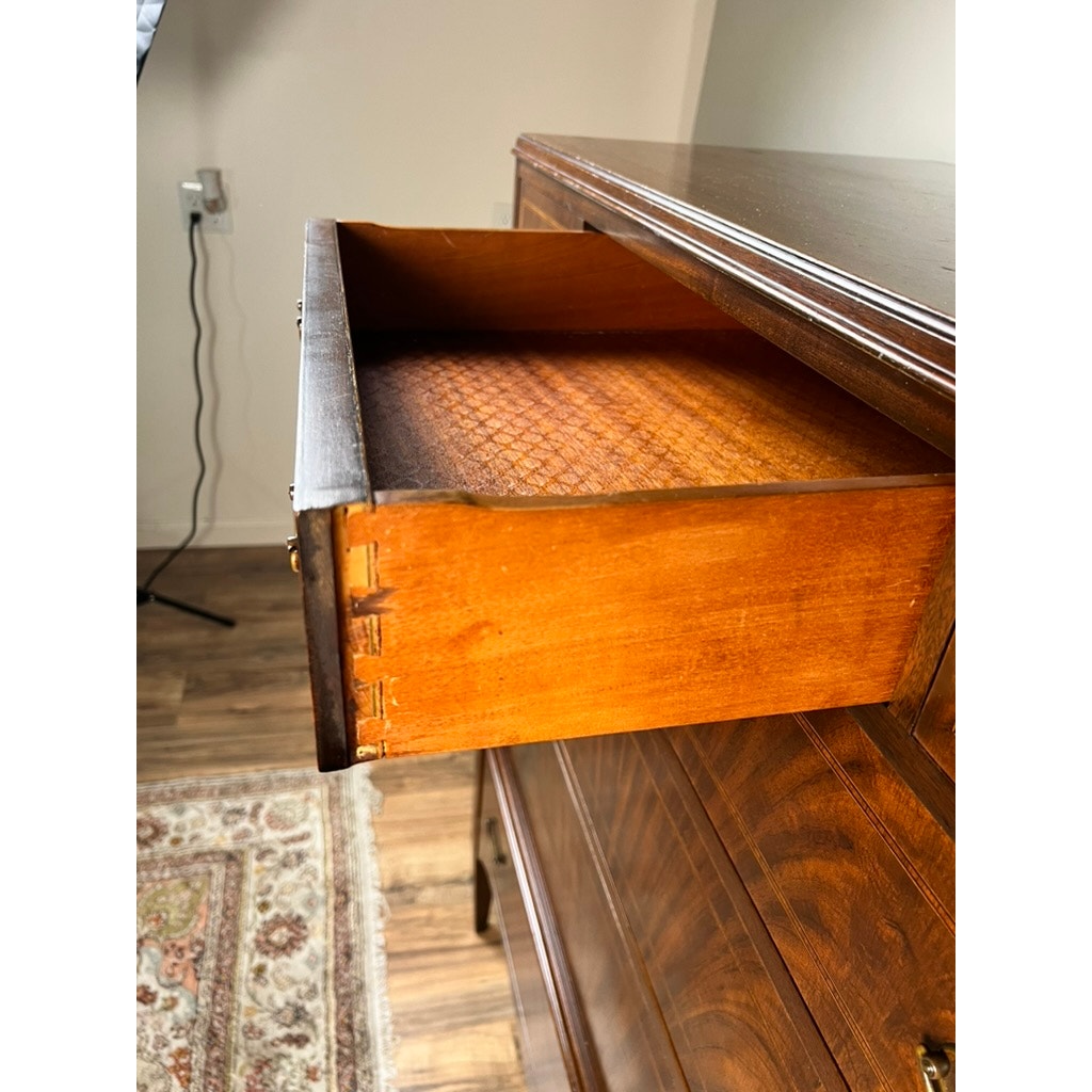 An open drawer of the Vintage Dutch Woodcraft Shops Federal Style Flame Mahogany Chest on Stand (c. 1945–1955) reveals classic dovetail joints. Part of a rug and a wall outlet can be seen to the left, complemented by wood flooring in the room.