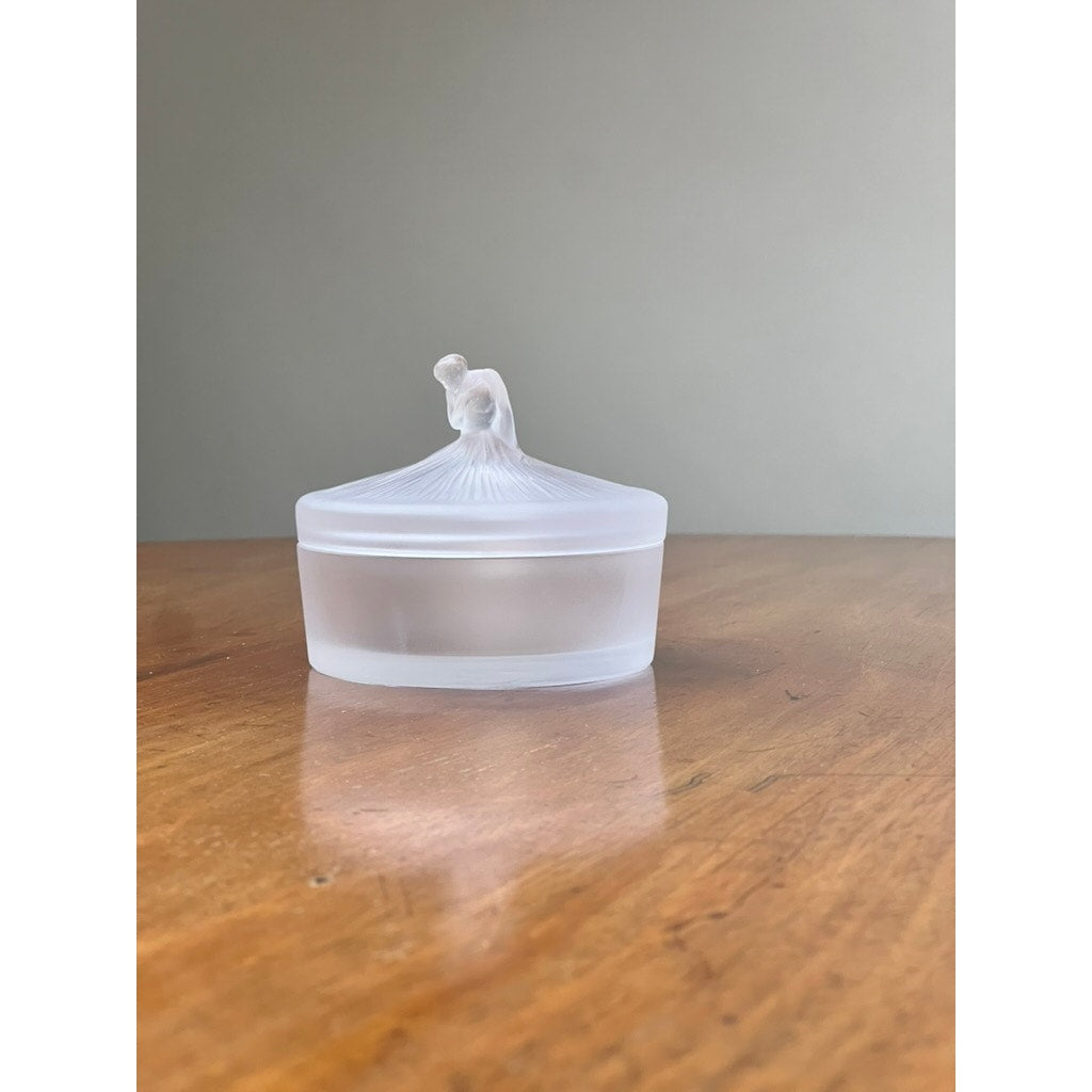 A Vintage Lalique France Ballerina Frosted Crystal Covered Powder Box by Lalique, featuring a round frosted glass container and sculpted ballerina lid, sits on a wooden surface against a plain, light background.