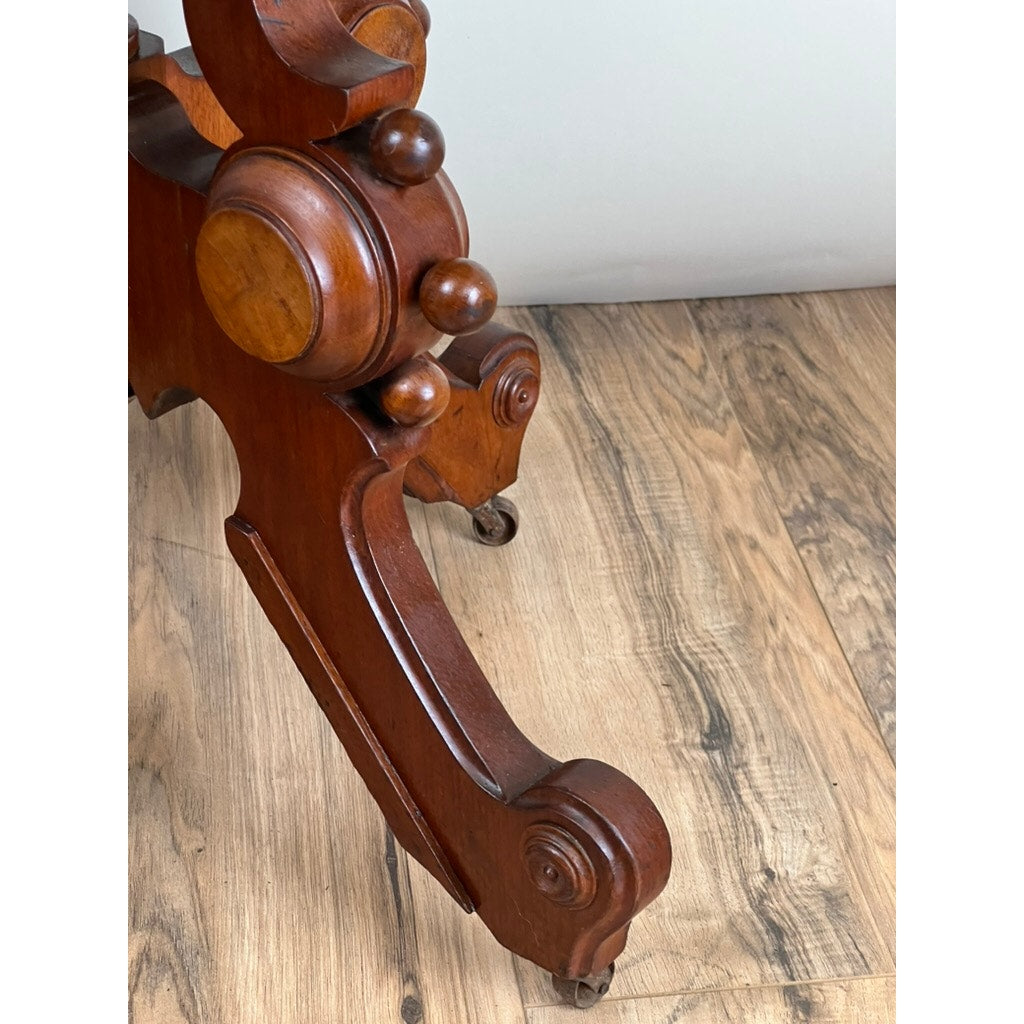 Close-up of an ornate, intricately carved leg with small wheels from an Antique Eastlake Victorian Walnut Marble Top Stand (late 19th c.), shown resting on a light brown wood floor. Brand: None/Unknown.