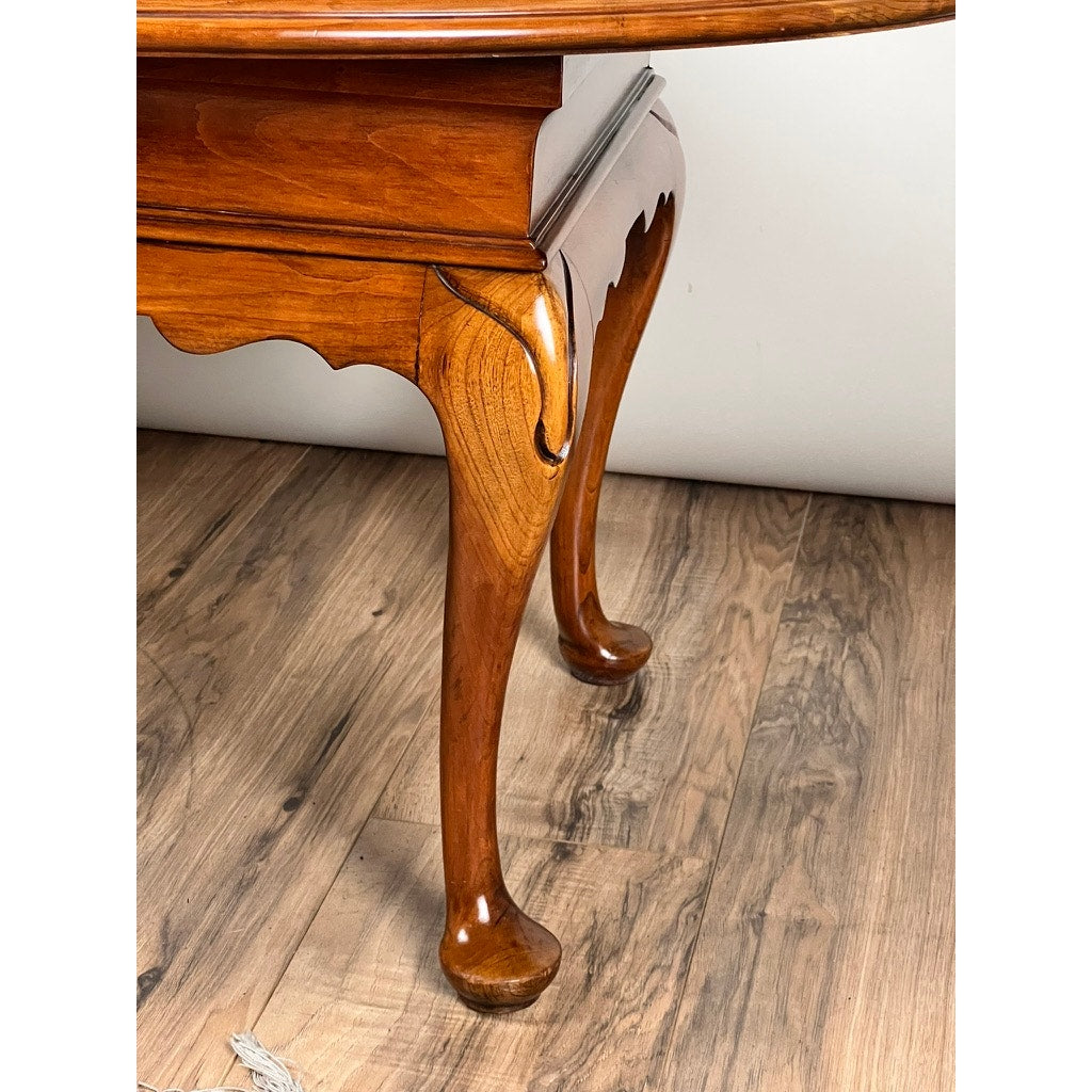 Close-up of a Vintage Solid Cherry Queen Anne Style Coffee Table leg, attributed to Pennsylvania House, with a cabriole design and claw-and-ball foot on wood-patterned flooring and a white wall in the background.