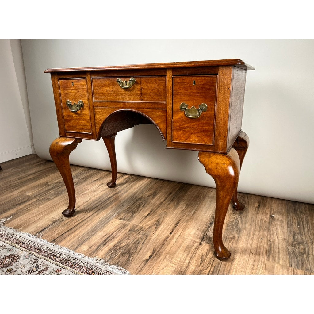 An Antique George III Queen Anne Mahogany Lowboy (late 18th century) with three drawers, brass handles, and cabriole legs is displayed on a wooden floor beside a patterned rug and a plain wall.
