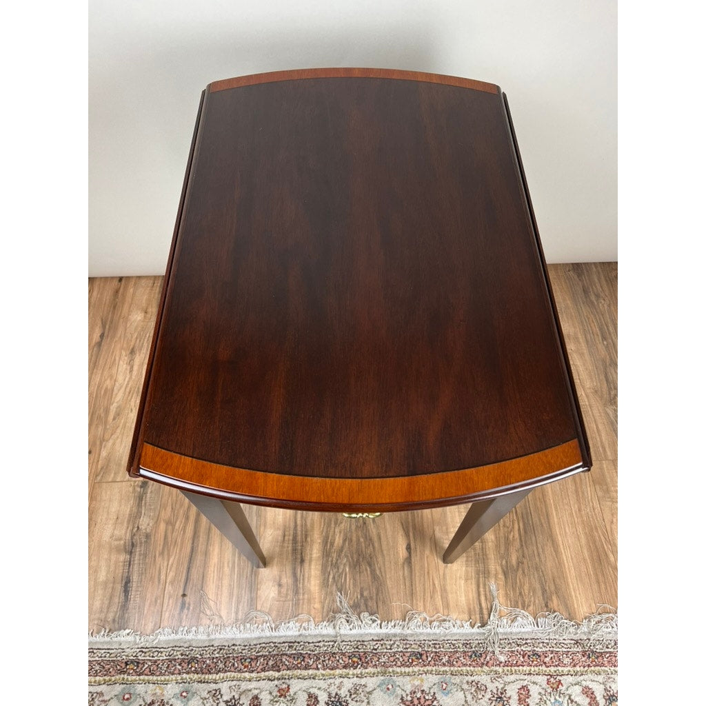 The Vintage Federal Style Edge Banded Mahogany End Table by Stickley, with a rectangular drop-leaf design and rounded edges, is shown from above on a wooden floor that's partly covered by a patterned rug.