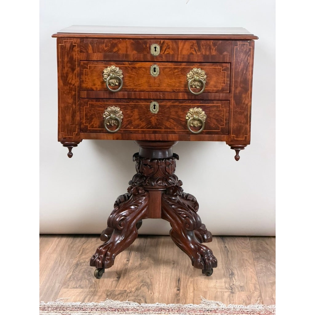 American Classical Figured Mahogany Work Table, circa 1830–1845, featuring two drawers with brass lion-head pulls and keyhole escutcheons, supported by a carved pedestal base with paw feet on casters.