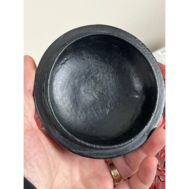 A hand holds the Antique Chinese Hand-Carved Cinnabar Lacquered Lidded Jar, probably Qianlong, with a round, shallow design and textured black lacquer. The background is blurred. Brand unknown.