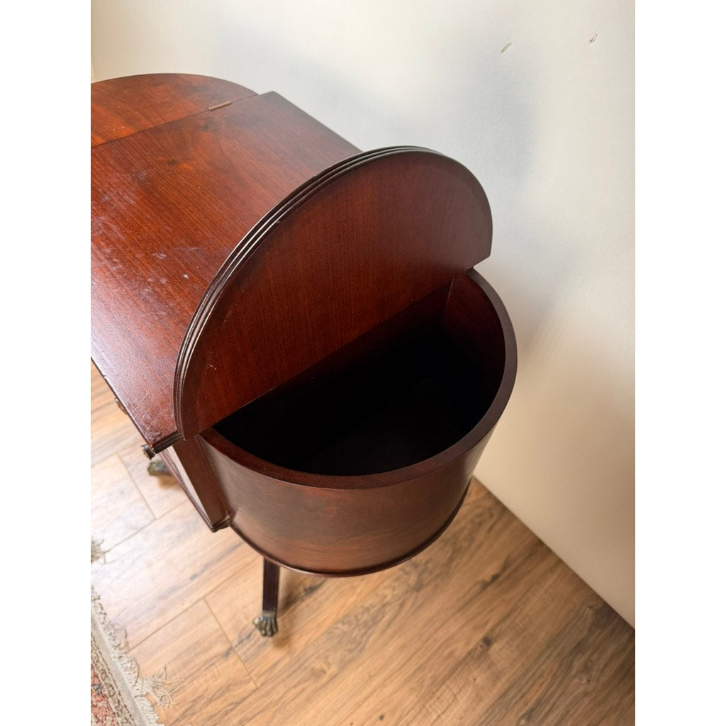 An Early 19th Century New York Classical Mahogany Astragal-End Work Table in the manner of Duncan Phyfe (c. 1815–1825) is shown with its semi-circular, hinged lid open to reveal storage space inside. Reminiscent of refined parlor furnishings from the era, this work table features the elegant design and craftsmanship characteristic of New York Classical mahogany pieces. The table is placed on a wooden floor beside a wall, with part of a rug visible nearby. Brand: None and/or Unknown.
