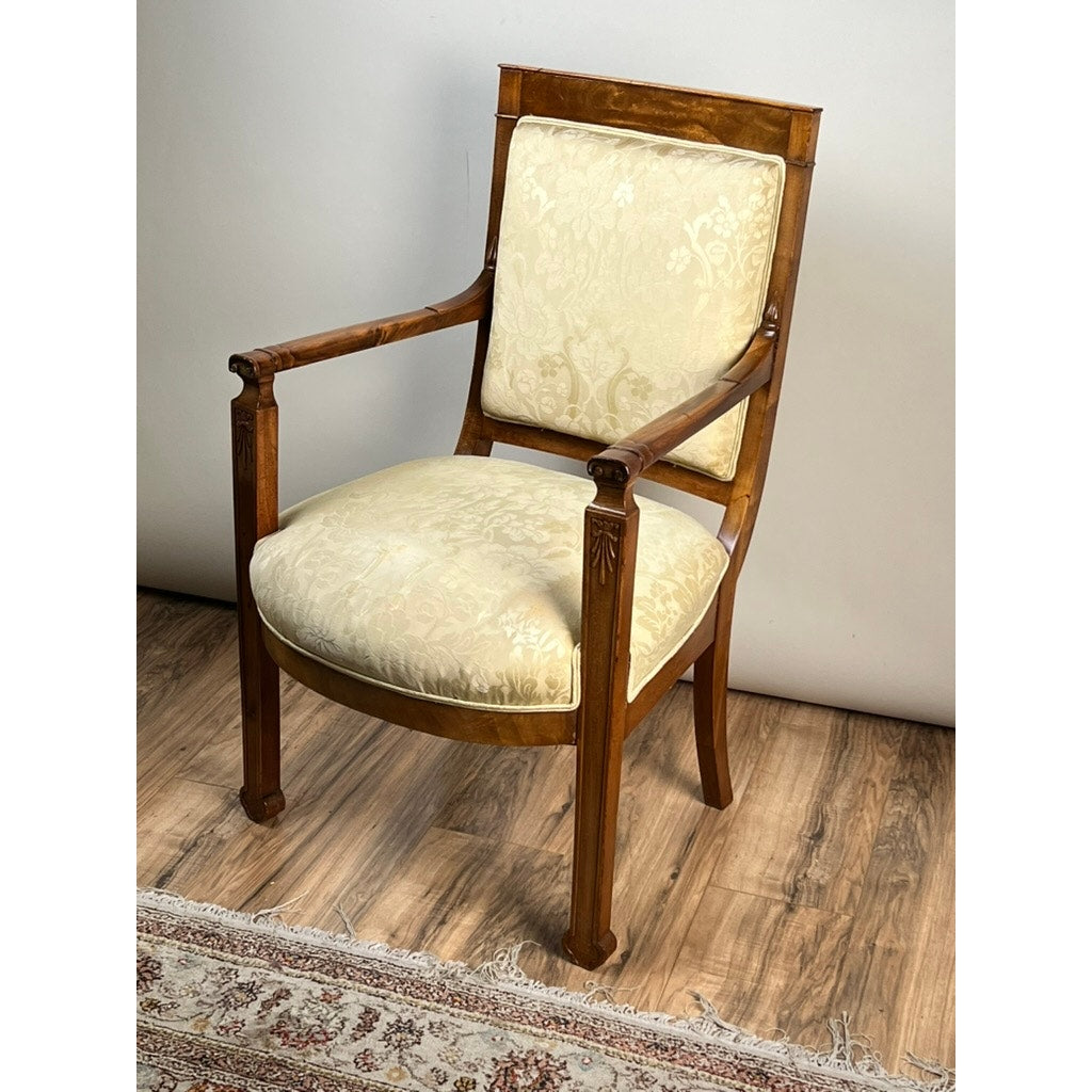An Antique French Directoire Style Fauteuil Open Arm Chair in Sapelle Mahogany, c. late 19th century, stands on a wooden floor beside a patterned rug and plain light wall, upholstered in cream-patterned fabric with carved details.