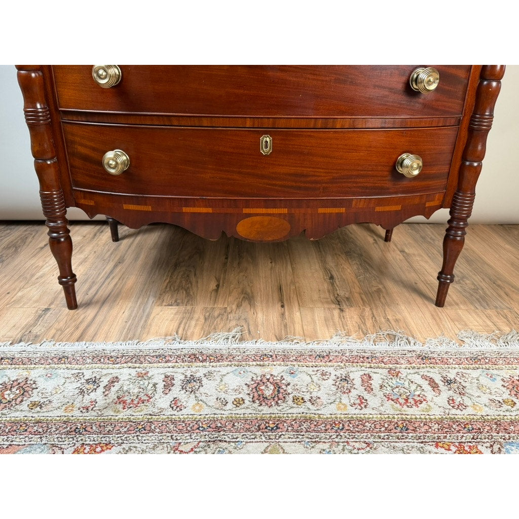 A close-up of an Early 19th Century New England Federal Mahogany Bow-Front Chest of Drawers with Backsplash, c. 1820–1840, featuring round brass handles, turned legs, and decorative inlay, standing on a wood floor next to a patterned fringed rug—an elegant example of early American craftsmanship.
