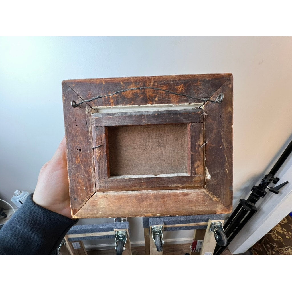 A hand holds the back of an Antique Oil on Canvas Small Format Painting of a Military Officer Escorting an Old Woman (c. 19th Century), showing its aged wooden frame, old nails, and hanging wire indoors near art supplies. No brand name.