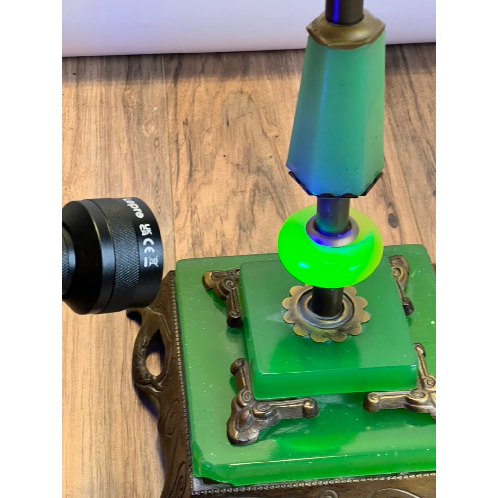 A close-up of an Antique Art Deco Uranium and Slag Glass Lamp c. 1920, its green glass glowing bright under a black UV flashlight, shown on a wooden surface. Brand: None/Unknown.