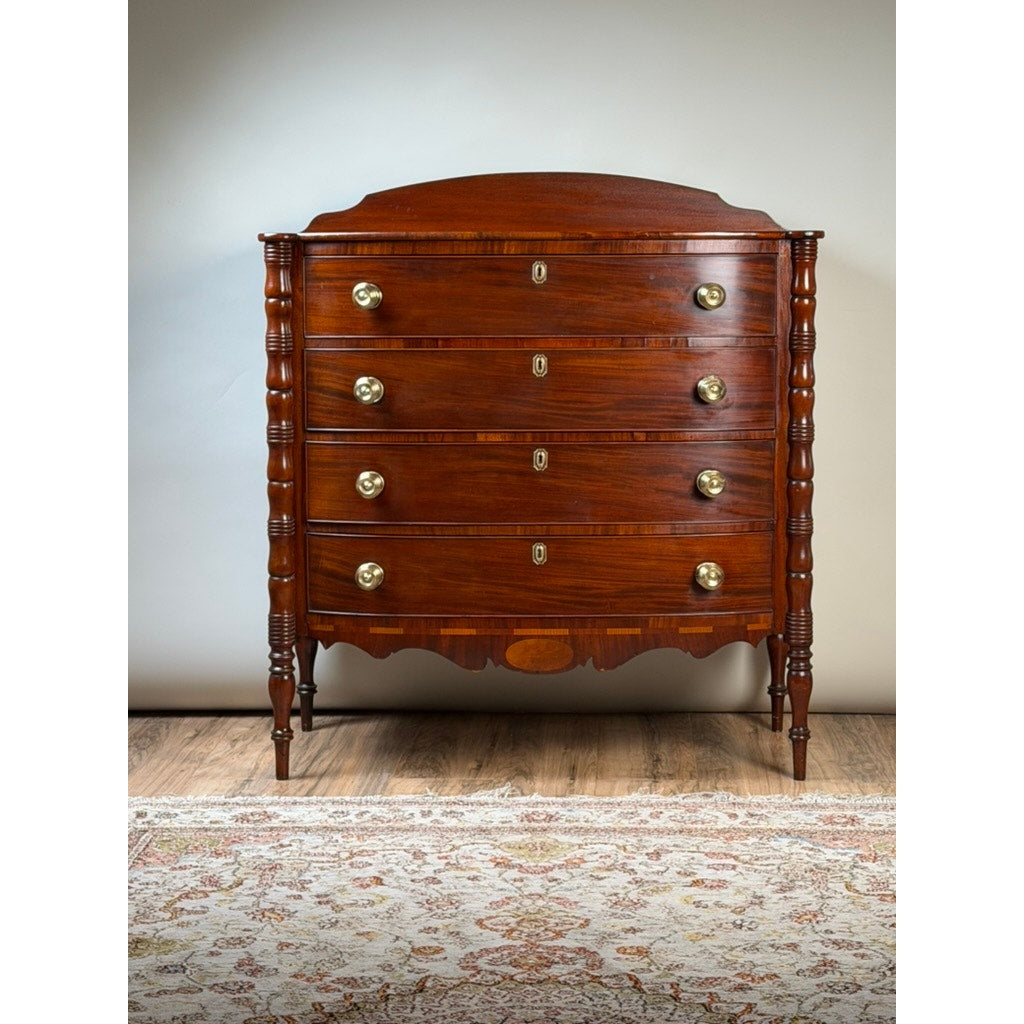 An Early 19th Century New England Federal mahogany bow-front chest of drawers with a backsplash, dating from circa 1820–1840, features four brass-handled drawers, turned legs, and a scalloped bottom, standing on a wooden floor with a patterned rug in front.