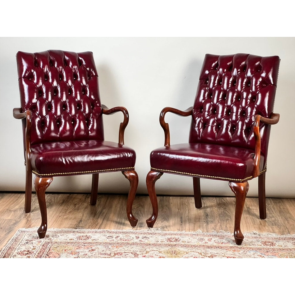 A Pair of Chippendale Style Tufted Burgundy Leather Library Armchairs, featuring wooden frames and cabriole legs, are placed side by side on a patterned rug against a plain wall.