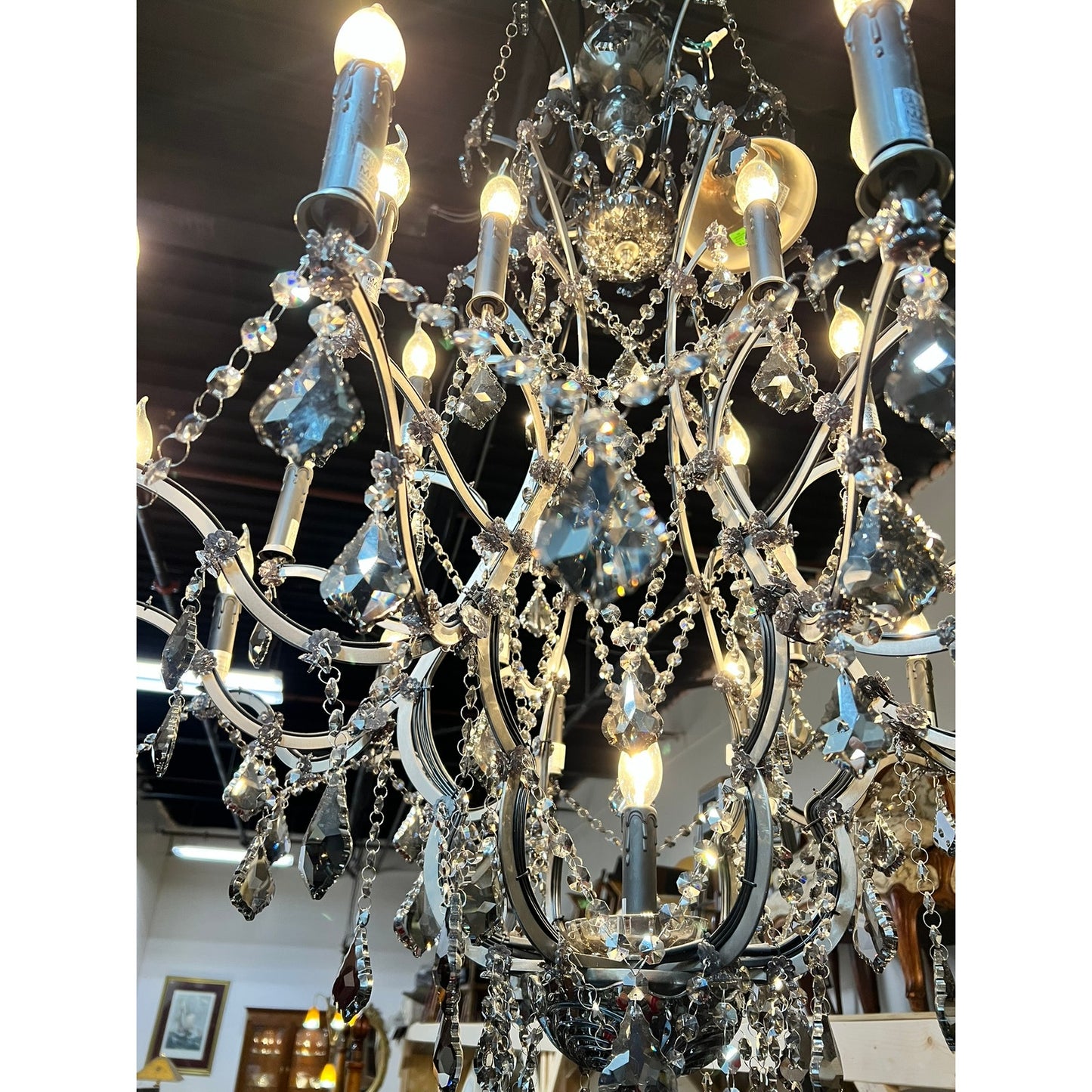 A close-up of the Large Vintage Smoke Crystal 24 Light Chandelier with glowing bulbs and tiered glass prisms, casting a sparkling light. A dark ceiling and framed art are visible in the background. Brand: None and/or Unknown.