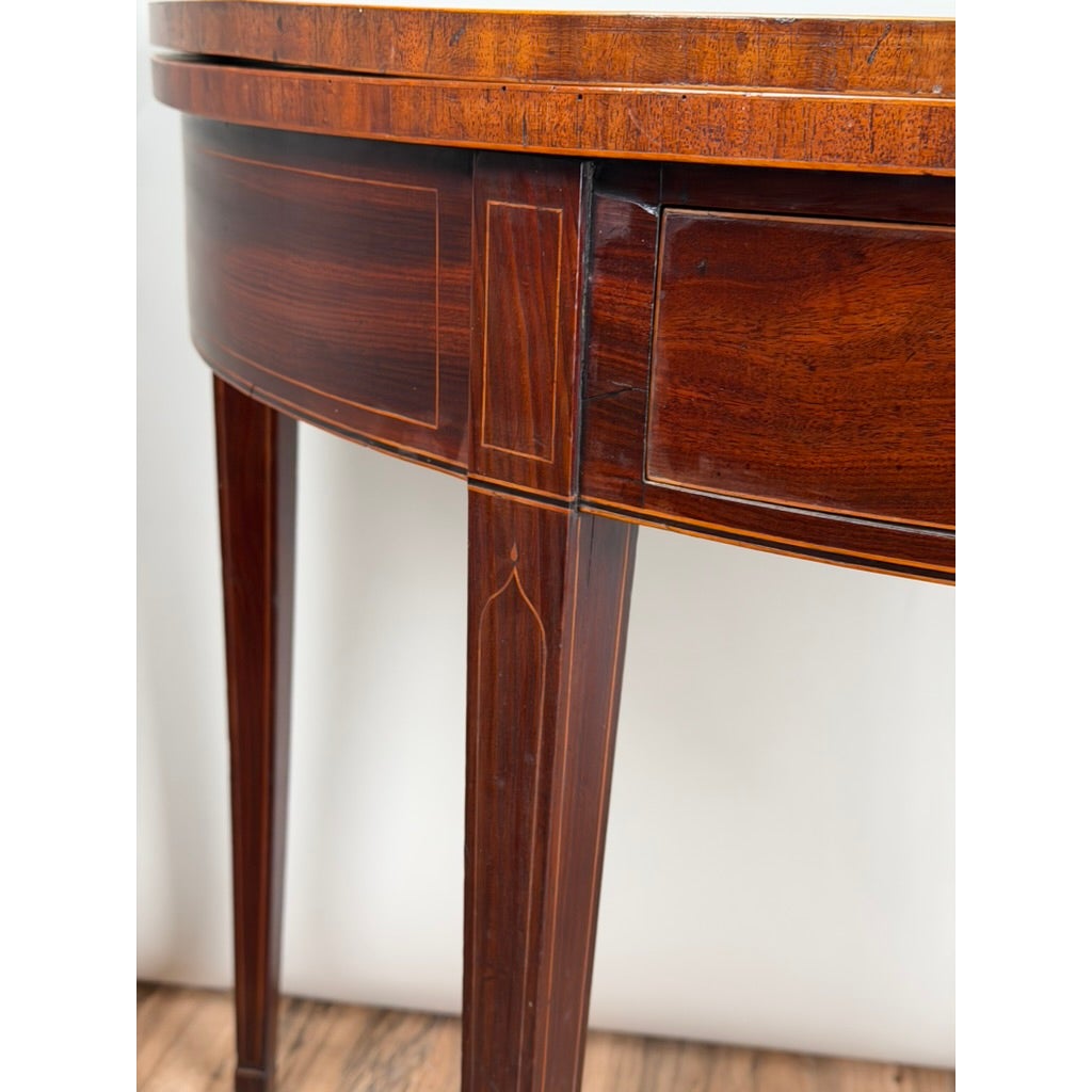 An English George III Mahogany and Satinwood-Inlaid Demilune Games Table featuring a semicircular wooden top, tapered legs, and inlaid light wood border details.
