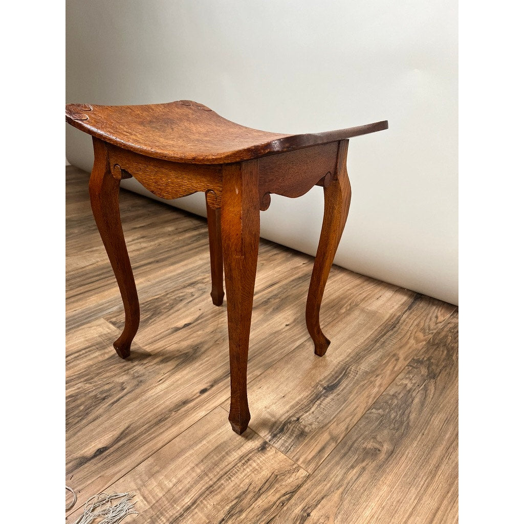 An Antique Arts and Crafts Saddle Shaped Footstool c. 1900, by an unknown maker, is displayed on a wooden floor against a plain white wall.