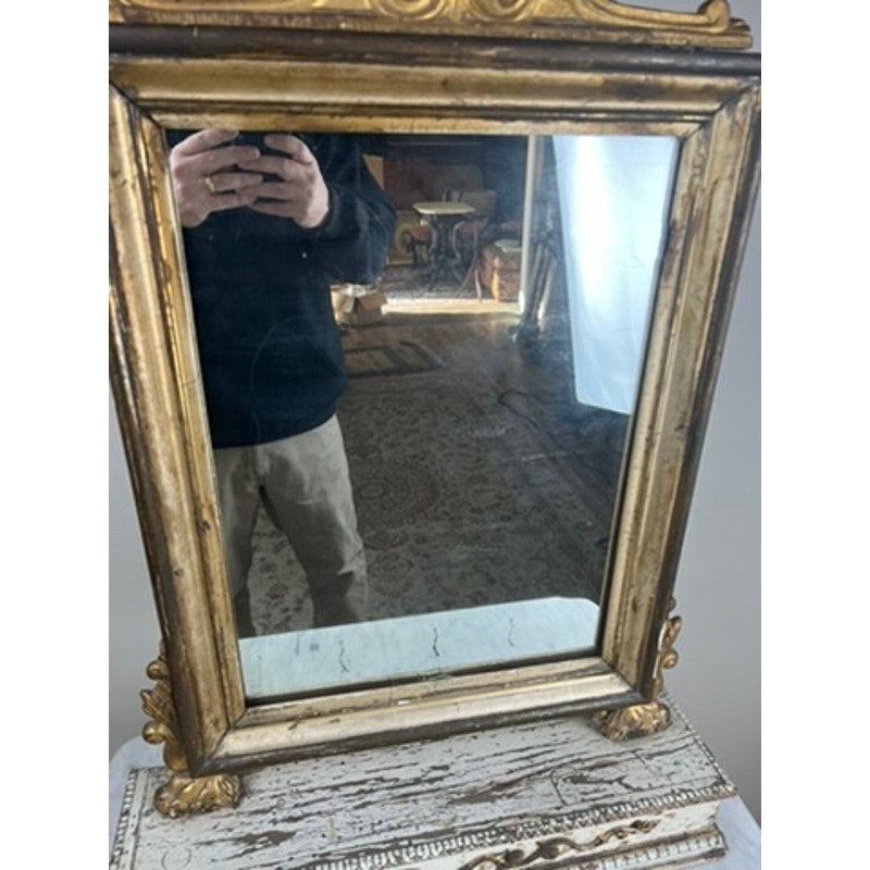 A person in a black top and beige pants is reflected in an Antique French Empire Gilt Wood Vanity or Table Mirror c. Mid 19th Century, resting on a distressed white wooden surface amid patterned carpet and antique furniture.