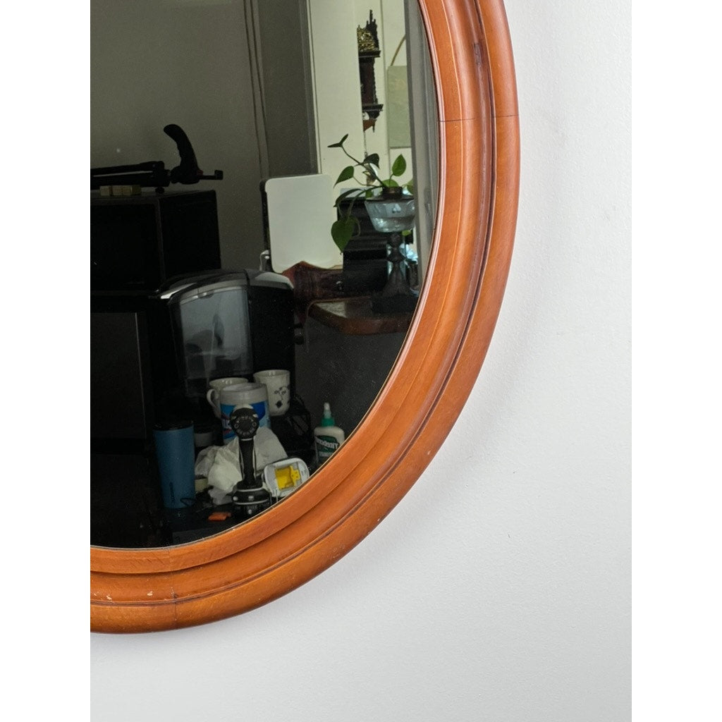 The Vintage Oval Maple Wall Mirror hangs on a white wall, reflecting a room with cups, bottles, a plant, and electronics. Brand: None/Unknown.