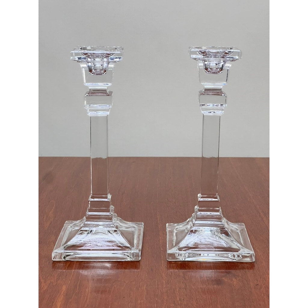 A Vintage Pair Tiffany & Co. Clear Crystal Candlesticks with square bases and geometric detailing sit side by side on a wooden surface, adding classic Tiffany & Co. charm to a plain, light-colored background.