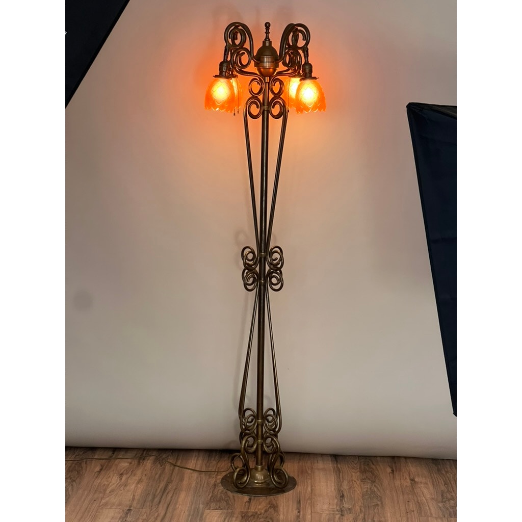 The Antique Art Nouveau Brass Floor Lamp, attributed to Jules Cayette and Charles Schneider c. 1920, features four lit orange glass shades and intricate scrollwork. It casts a warm glow on the wooden floor against a plain wall.