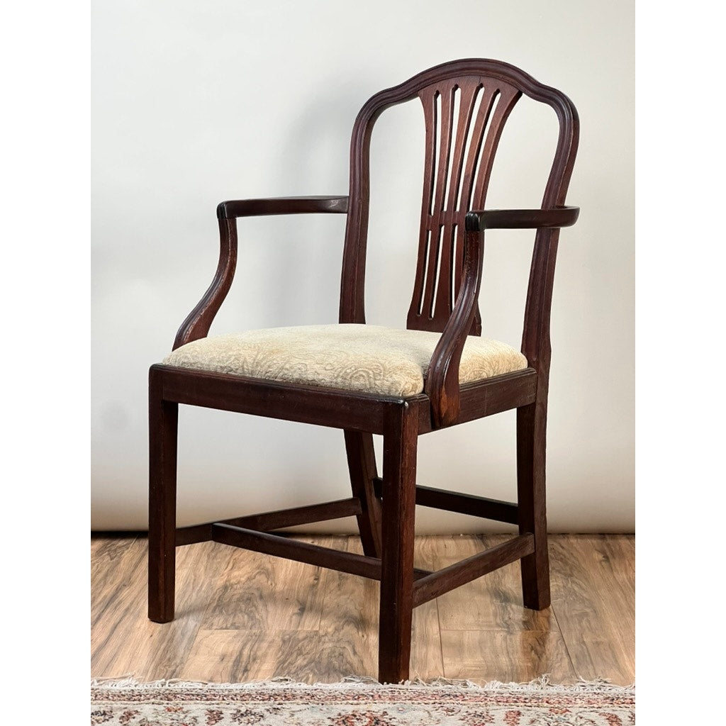 An Antique English George III Mahogany Hepplewhite Armchair (Early 19th Century) with an ornate curved backrest, vertical slats, and a light patterned fabric seat stands on a wooden floor beside a patterned rug and plain light wall.
