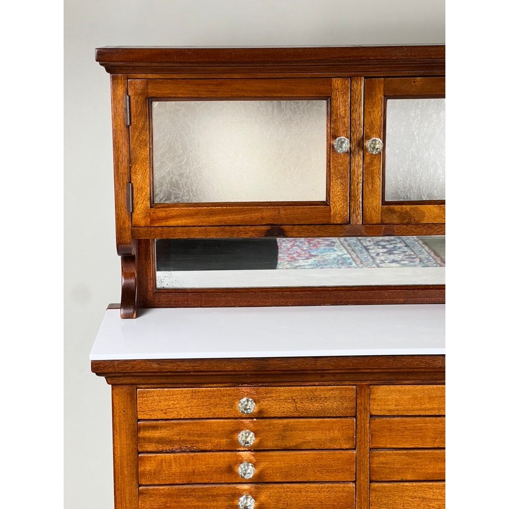 A close-up of an Antique Mahogany Multi-drawer Dental Cabinet by The American Cabinet Co. (early 20th c.) shows textured glass doors, decorative glass knobs, a mirrored back reflecting a patterned rug, white countertop, and wood drawers.