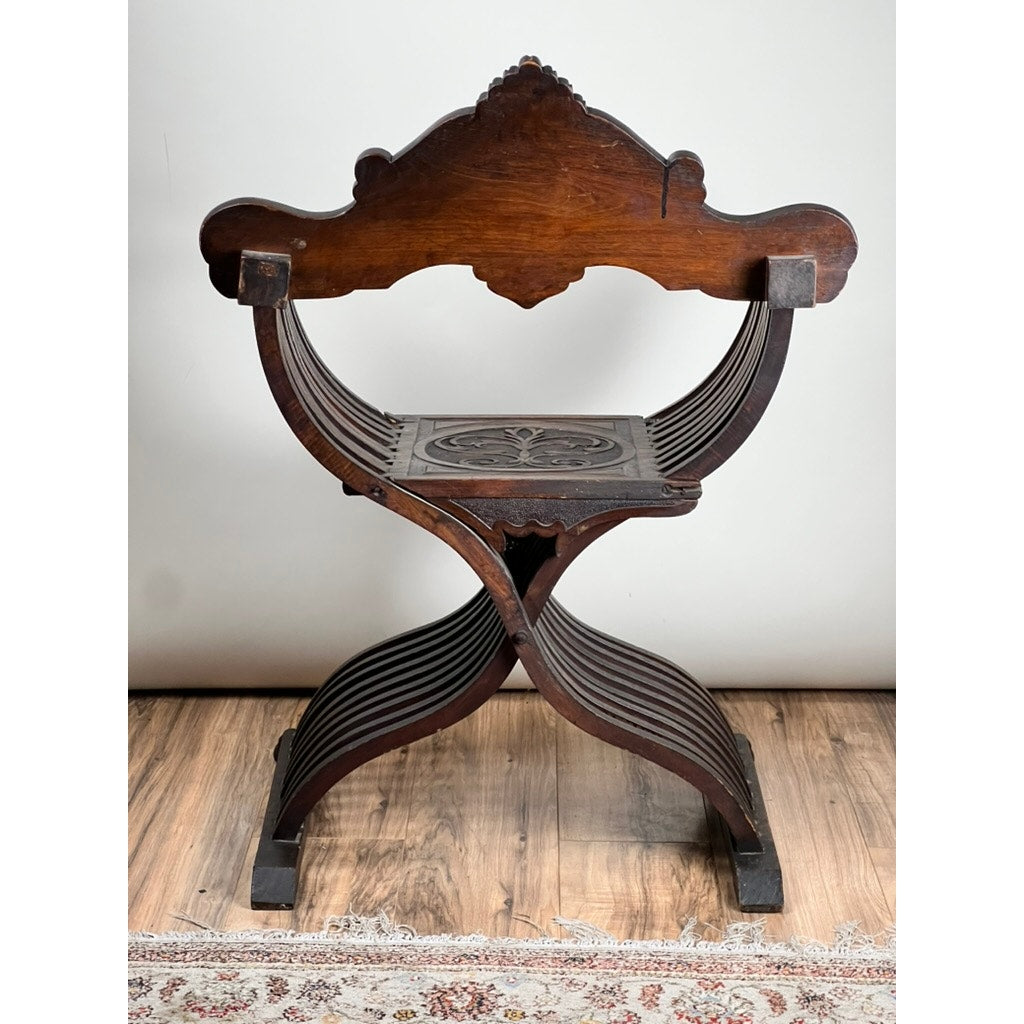 An Antique Italian Renaissance Walnut Savonarola Chair (late 19th century) with ornate carvings, curved X-shaped legs, and a decorative backrest stands on a wood floor by a patterned rug against a light wall—a fine historic accent.