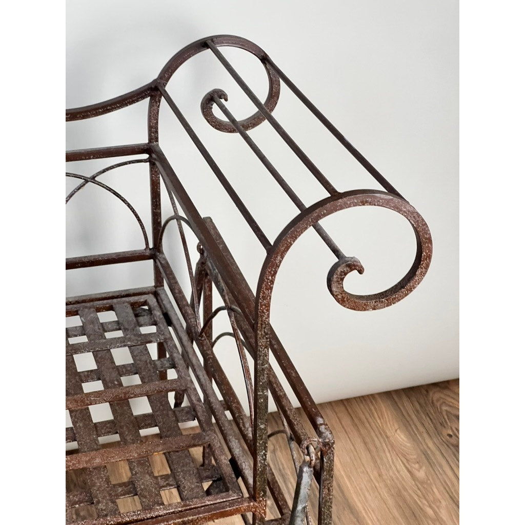 Close-up of a Vintage Regency Style Wrought Iron Porch Glider showcasing ornate scrollwork armrest and lattice seat, evoking a classic garden feel, set against a white wall and wooden floor. Brand unknown.