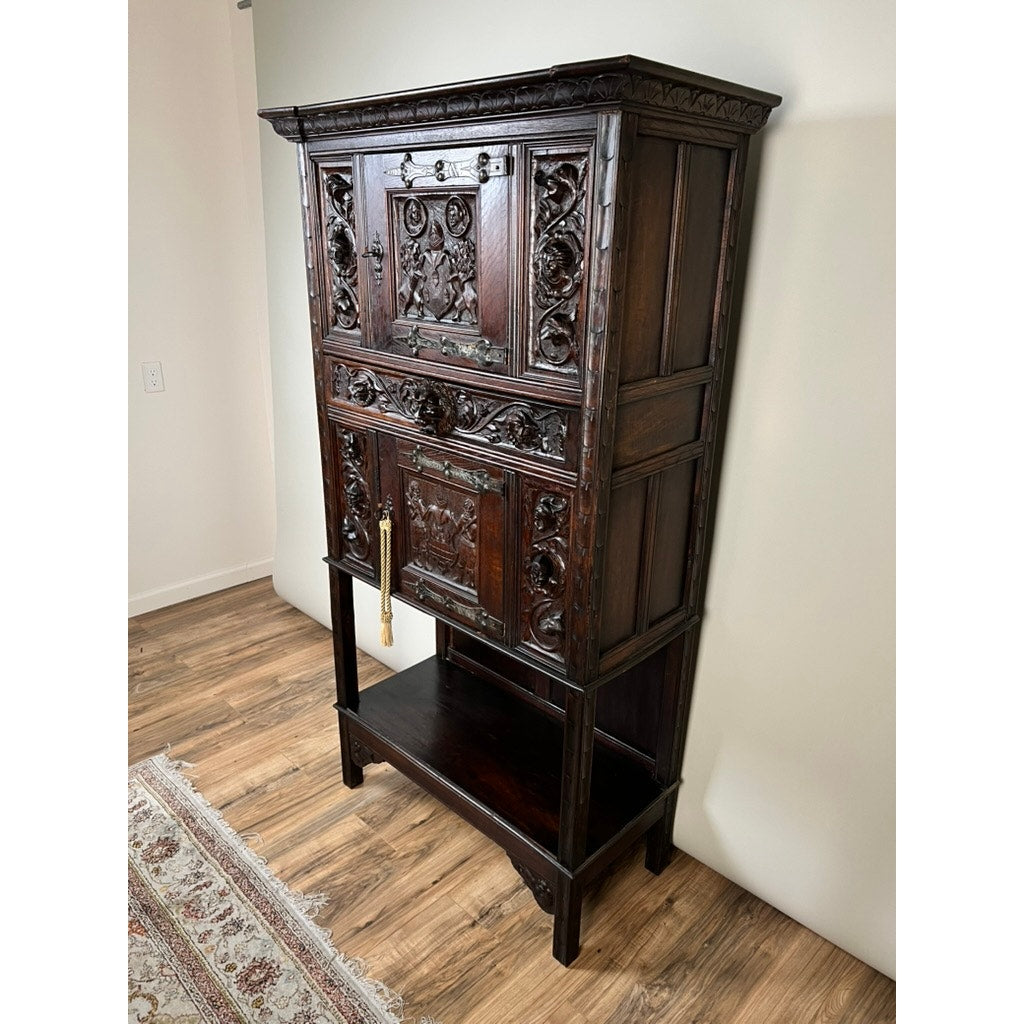 Antique 19th-century French Breton-style carved oak cabinet featuring deeply carved knights, with two closed doors, a drop-front desk section, and an open lower shelf. The piece stands against a light wall on a wooden floor near a rug.