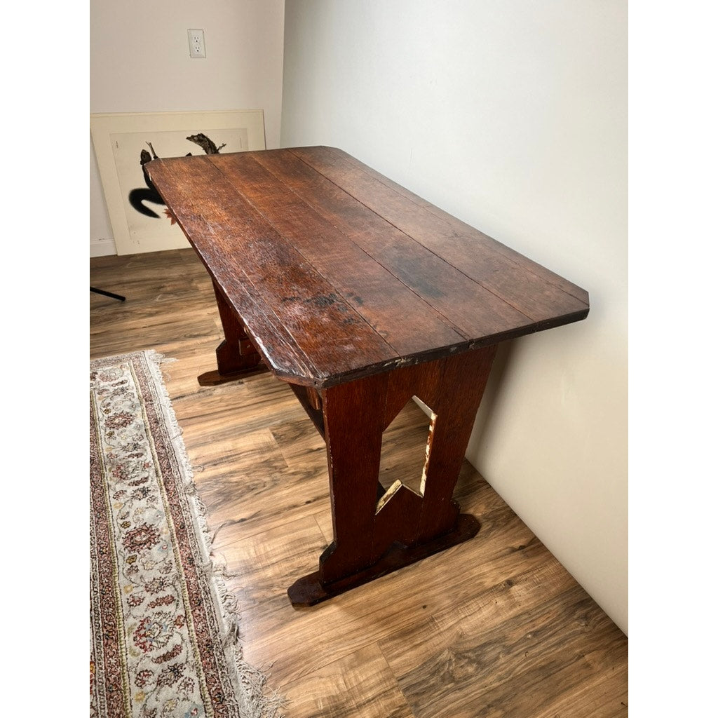An Antique Rustic Italian Fratino Trestle Table (late 19th century) with sturdy, angled legs and a decorative cutout stands on a wood floor by a patterned rug, positioned against a white wall. Brand unknown.