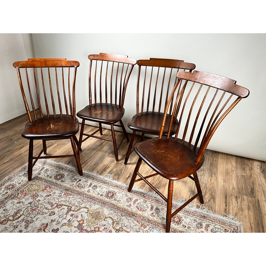 A set of four early 19th-century New Hampshire Step Down Windsor chairs by JR Hunt, showing visible wear, stands on a patterned rug over a wood floor against a light wall.