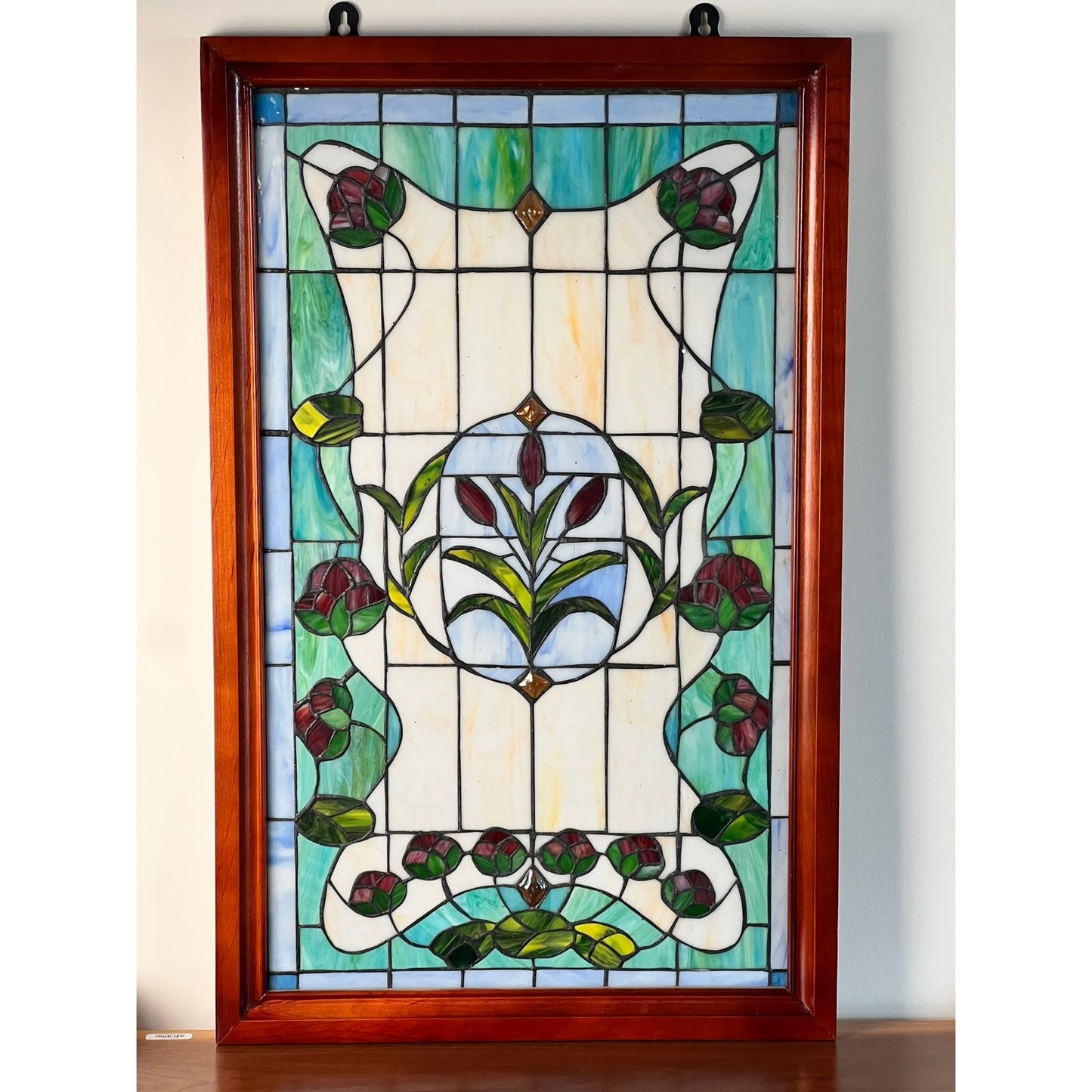 The Vintage Stained Glass Wall Hanging 32x20 showcases a floral and leaf Art Nouveau design in green, red, and blue with a central circular motif and decorative borders. Displayed on a wooden surface against a white wall. Brand unknown.