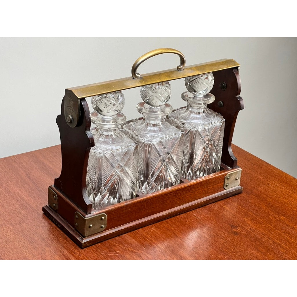 A Betjemann's Antique English Edwardian cut crystal three-bottle tantalus (early 20th century) with matching stoppers sits on a polished wooden table.