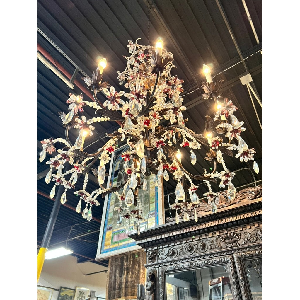 An Antique Late 19th Century French Rococo Tole Amethyst and Crystal Ten Light Chandelier hangs from a dark ceiling above an intricately carved wooden cabinet, adorned with amethyst accents and sparkling crystal prisms.