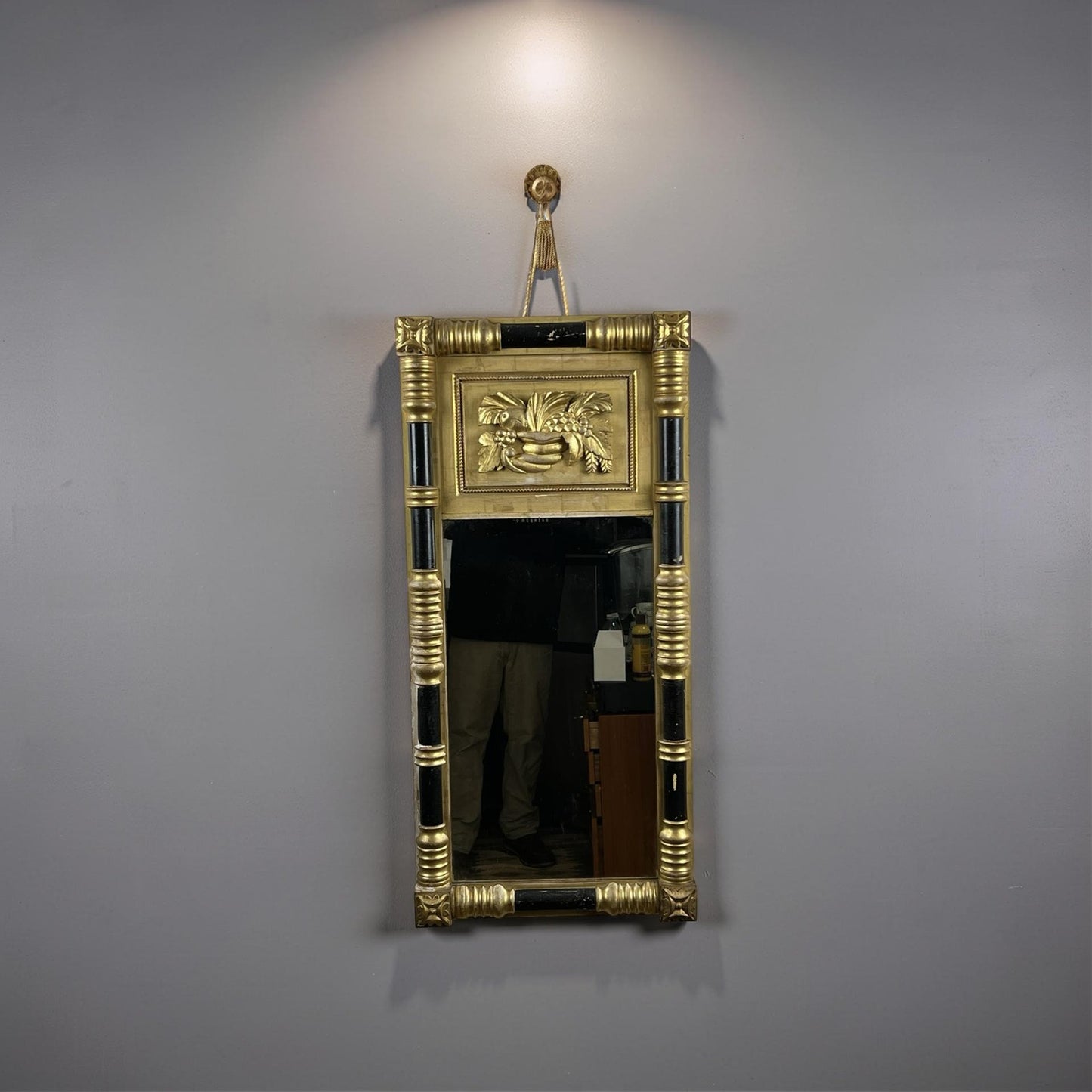 An Antique Federal Period Giltwood Mirror with Rosette Corners and Cornucopia c. 1830 hangs on a gray wall under a spotlight; a faint reflection of a person's lower body is visible in the mirror’s glass.