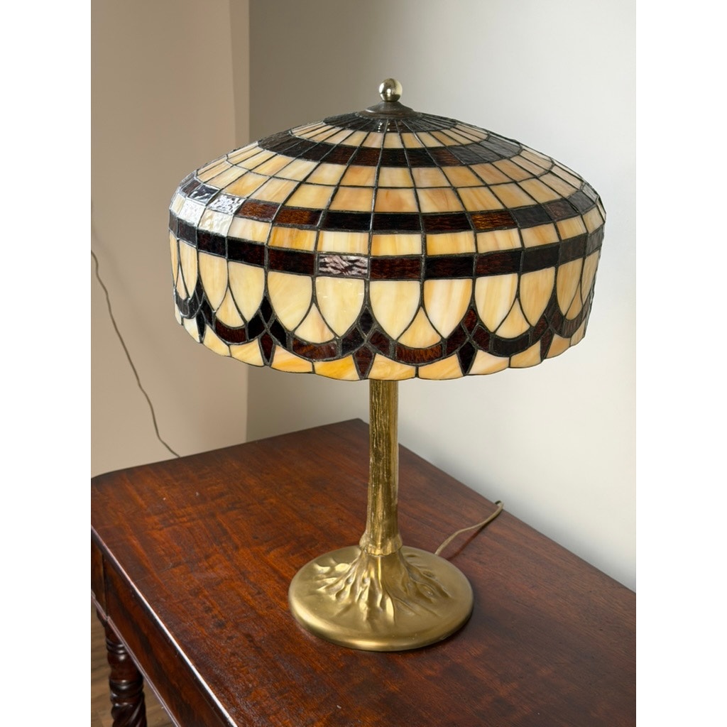 A Pearl City Antiques Vintage Tiffany Revival Leaded Stained Glass Table Lamp with a cast brass tree-form base rests on a polished wooden table near a light-colored wall. The lamp’s leaded stained glass shade displays an elegant pattern in cream, brown, and black geometric motifs.