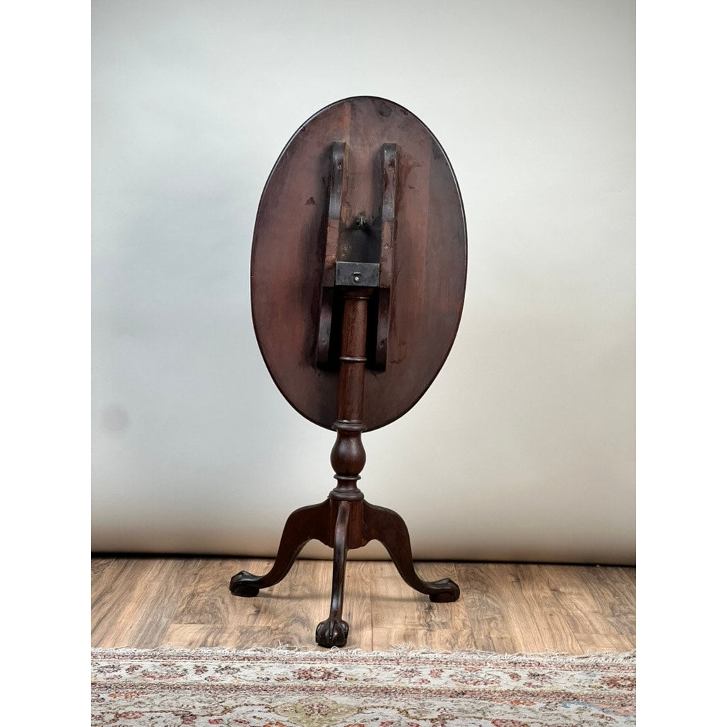 An Antique George III Tilt-top Tea Table from the early 19th century by Pearl City Antiques, distinguished by its oval tilt-top and gracefully supported on a tripod base with claw feet, stands upright on a wooden floor with part of a rug visible in front.