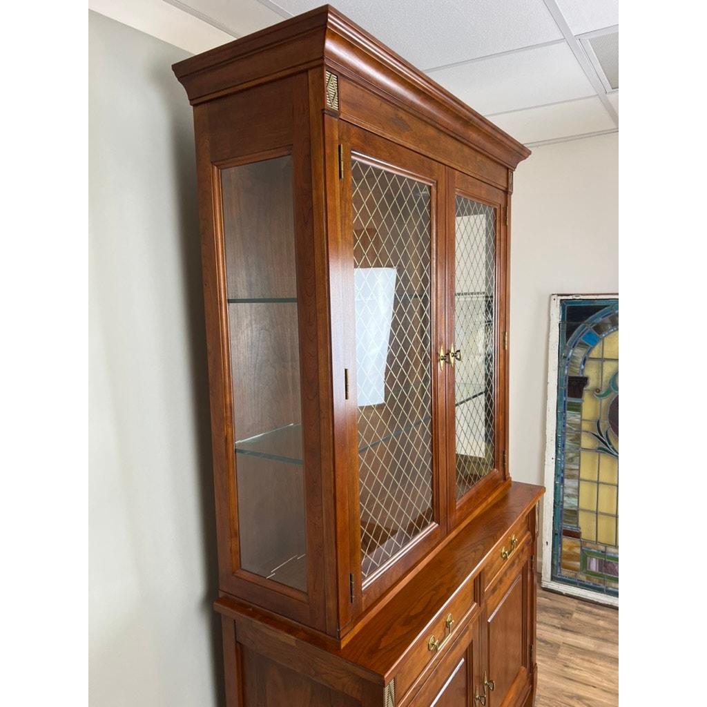 The Vintage William IV Style Cherry Glass Door Bookcase by Stickley features glass panel doors with brass mesh inserts and interior glass shelves, shown against a white wall and wood floor, with a stained glass piece nearby.