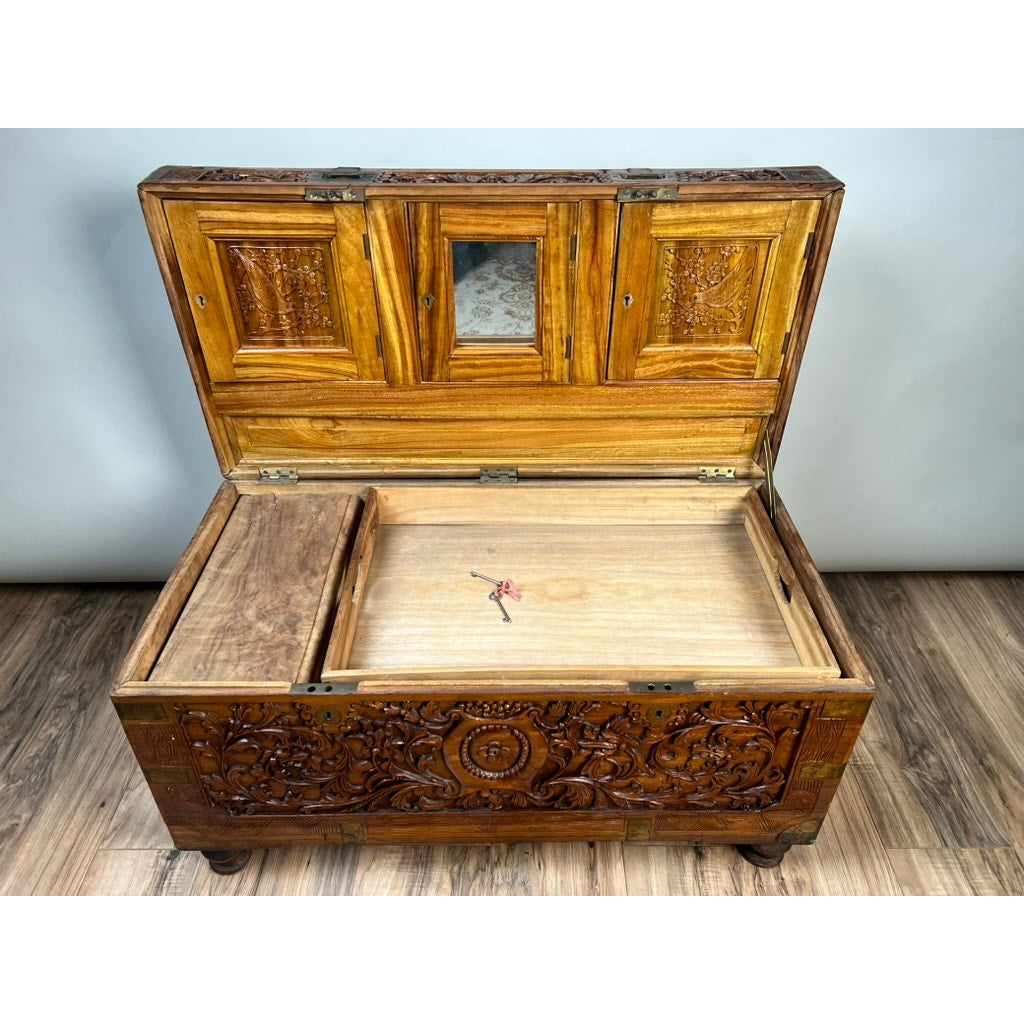 A Chinese Export Carved Camphor Wood Dowry Chest, adorned with intricate hand-carvings, stands gracefully on a wooden floor. The open lid reveals multiple storage compartments, a sliding wooden tray, and a small mirror set inside the lid.