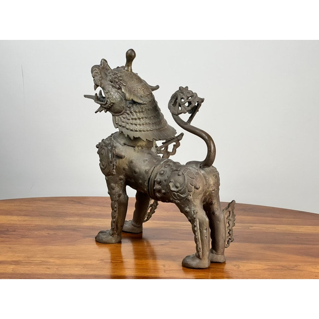 The Vintage 17” Cast Brass Pixiu Tibetan Lion c. 1960 statue, featuring scales and intricate details, stands on polished wood in front of a white background, reflecting classic Tibetan Chinese decor. Brand: None/Unknown.