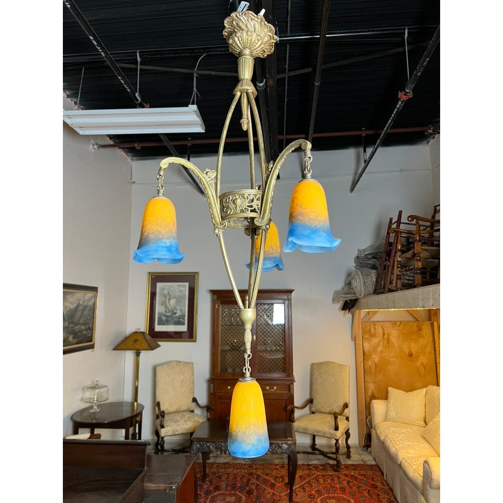 A Vintage Muller Frères Lunéville French brass chandelier with four Art Glass shades by Muller Frères hangs in a vintage-furnished room with chairs, a cabinet, and framed art on the walls.