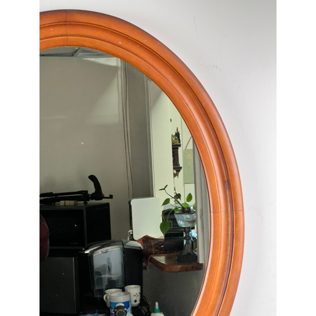 A Vintage Oval Maple Wall Mirror reflects a room scene with shelves, a plant in a glass vase, electronics, and stacked cups by a coffee machine. Brand: None or unknown.
