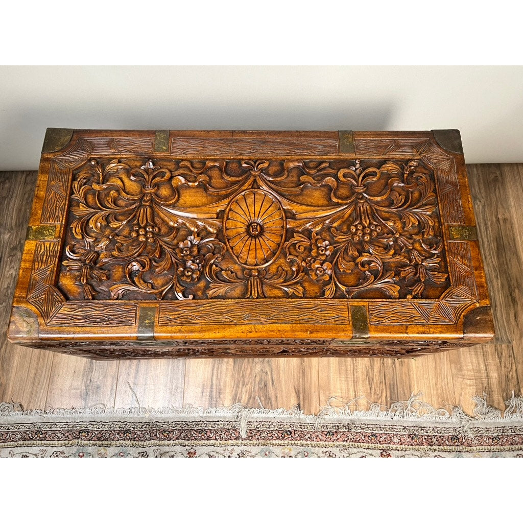The Chinese Export Carved Camphor Wood Dowry Chest, featuring intricate floral carvings on the lid, brass corner accents, and a central oval design, sits elegantly on a hardwood floor near a fringed rug.