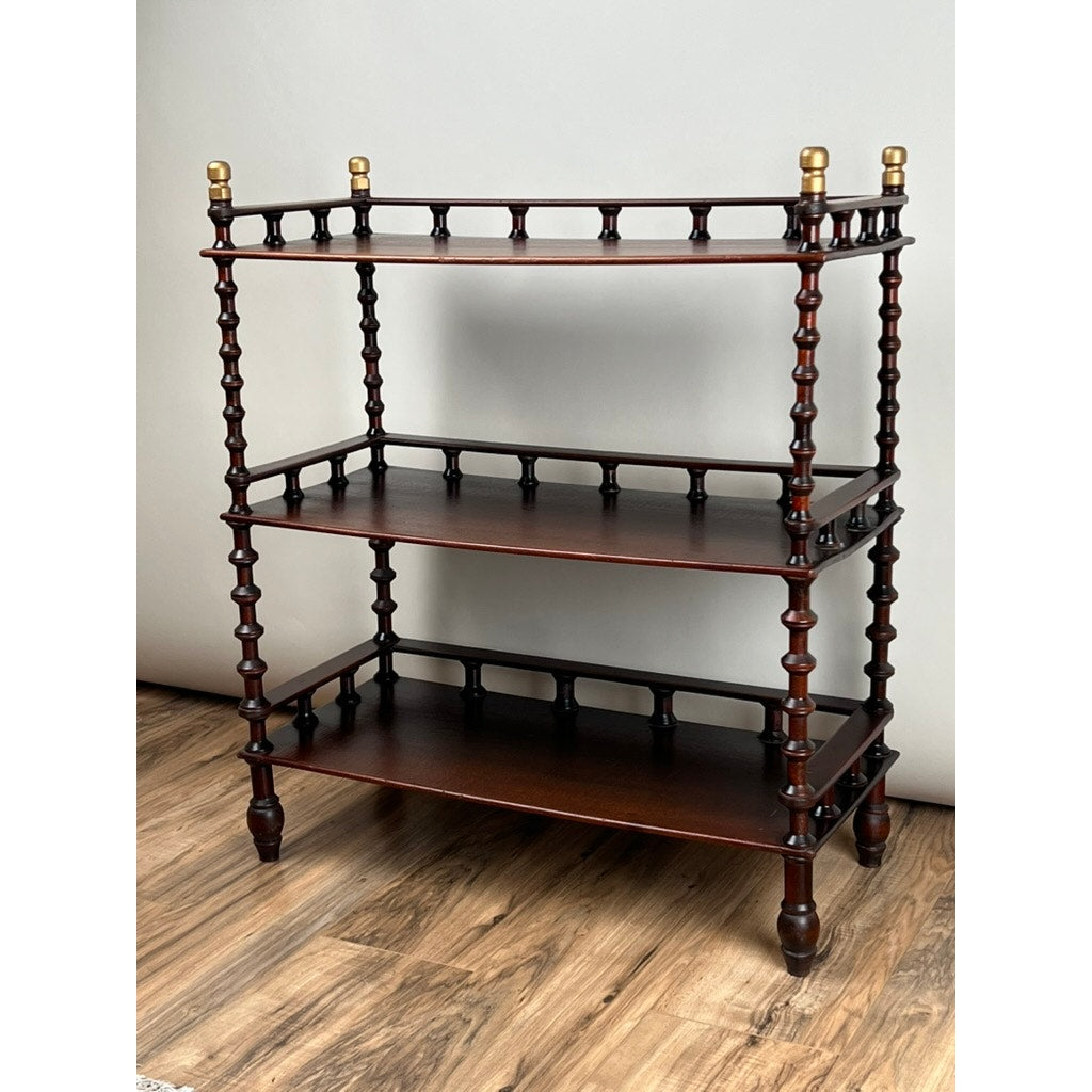 The Vintage Bobbin-turned, Three Shelf Wooden étagère or Whatnot features turned legs, spindle gallery accents, and brass finials on top corners. Its dark-finished shelves create a striking contrast against light backgrounds on a wooden floor.