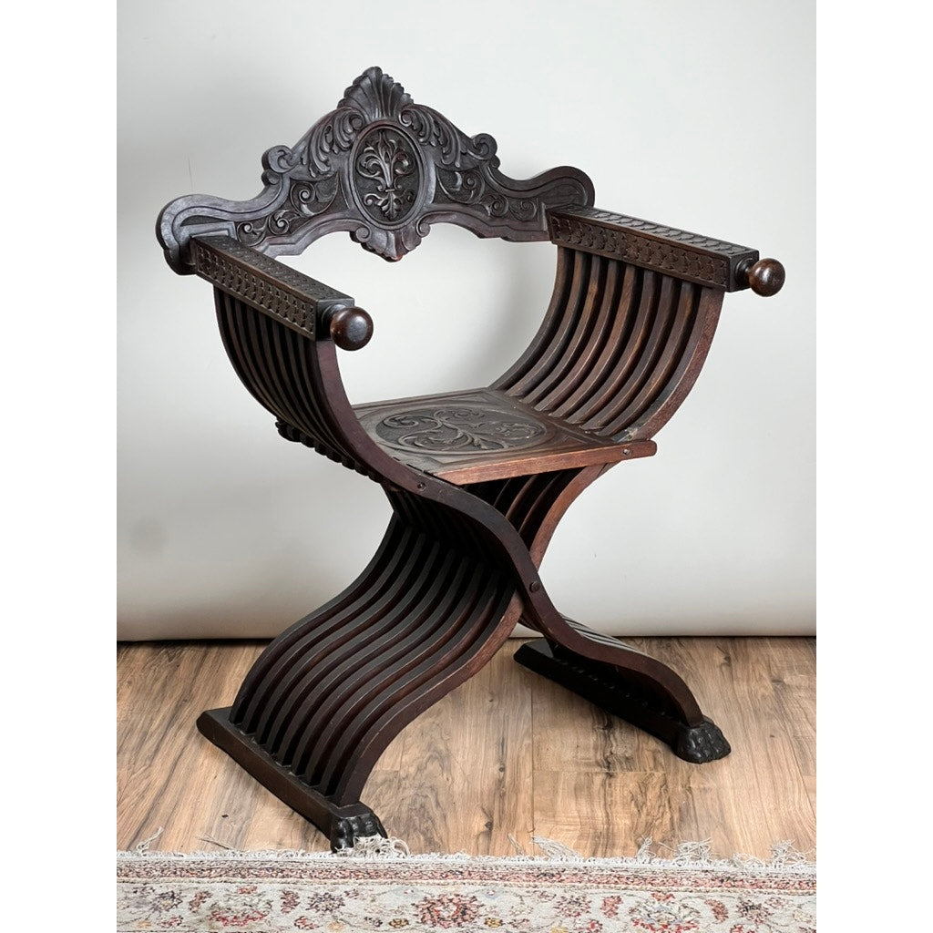 Antique Italian Renaissance Walnut Savonarola Chair (late 19th c.) with curved X-shaped legs and ornate carvings on backrest and arms, shown on a wooden floor next to a patterned rug and plain wall. Brand: Unknown.