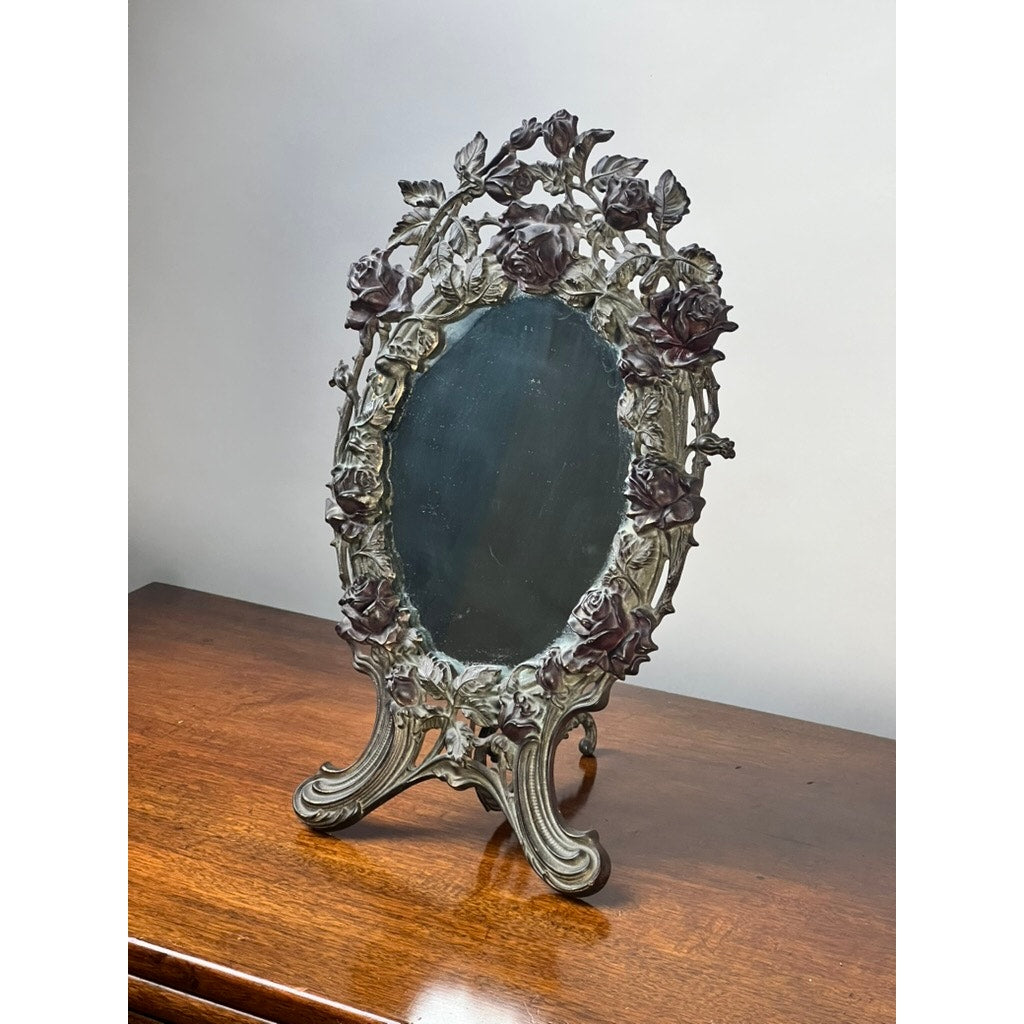 The Antique Art Nouveau Silvered Cast Brass Rose Vanity Dressing Mirror, dated 1909, with an ornate oval frame of intricate rose designs, stands on a polished wooden surface against a plain light-colored wall.