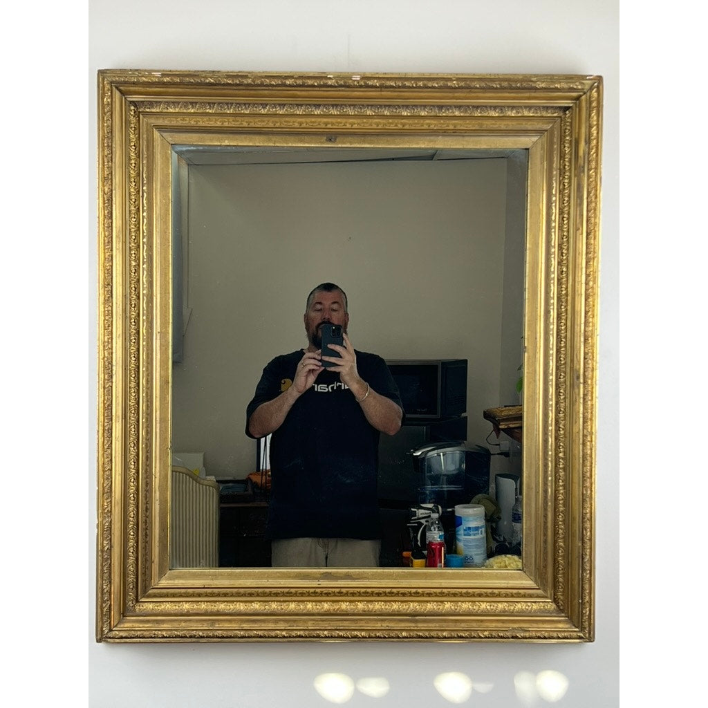 A person takes a selfie in a Fine Antique 19th Century Gilt Wood Mirror (39 x 34) mounted on the wall, with a TV, furniture, and household items in the background. The phone is visible, and light spots appear below the mirror.