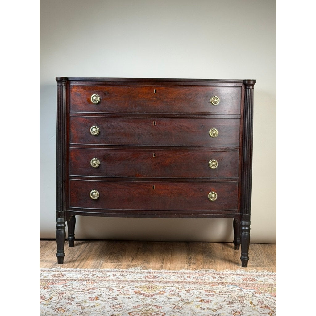 An Early 19th Century New England Federal Mahogany Bow-Front Chest of Drawers, dating from circa 1810–1825, with four drawers, brass round handles, and elegant tapered legs stands on a hardwood floor with a patterned rug in front.